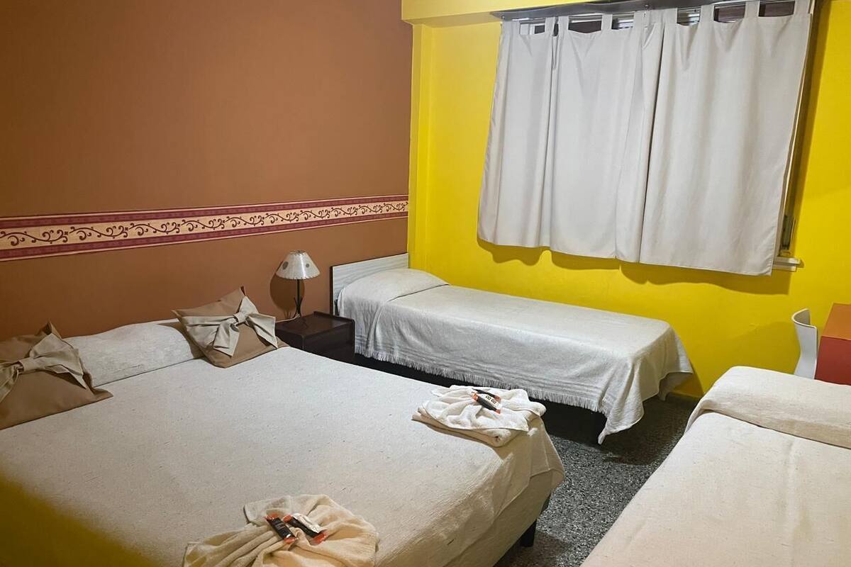 Habitación de hotel con dos camas individuales, una mesita, una lámpara y un ventanal con cortinas.