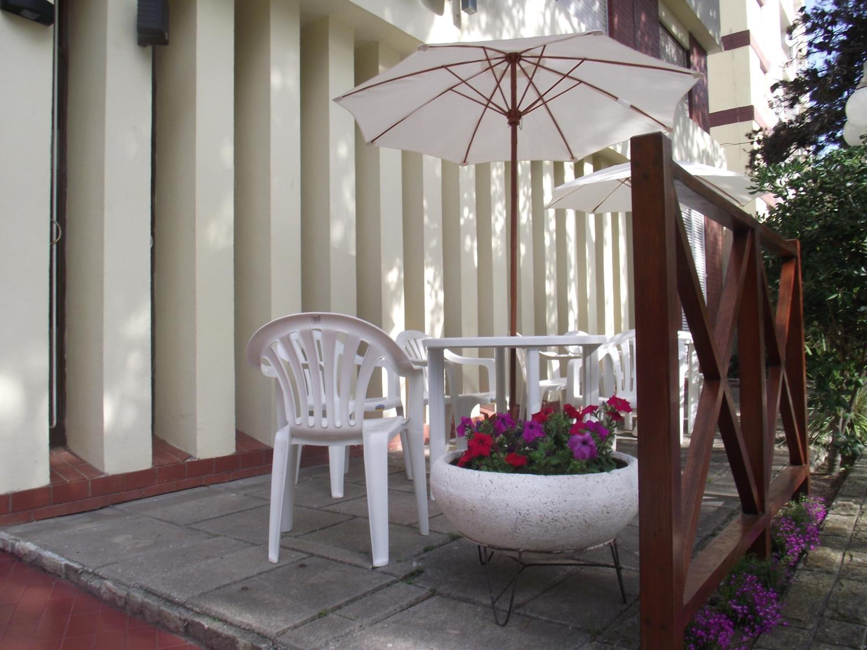 A patio with white plastic chairs, a white umbrella, and a concrete planter with flowers.