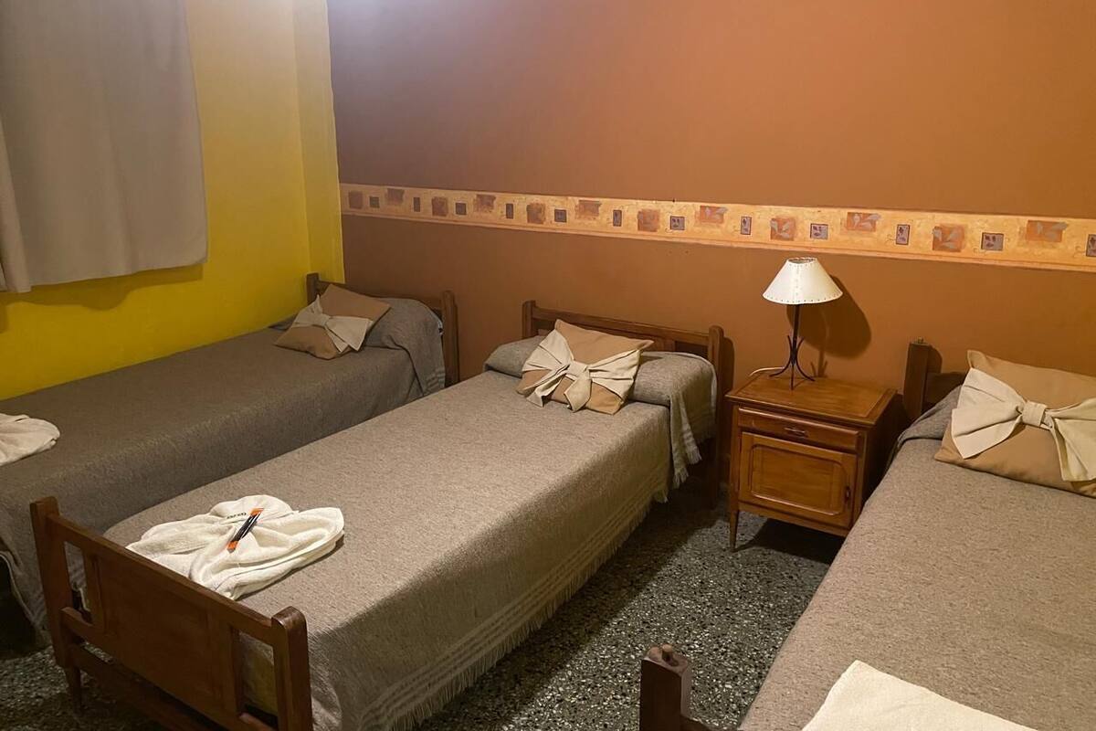 Una habitación de hotel con tres camas individuales, una mesita de noche con lámpara y una pequeña mesa.