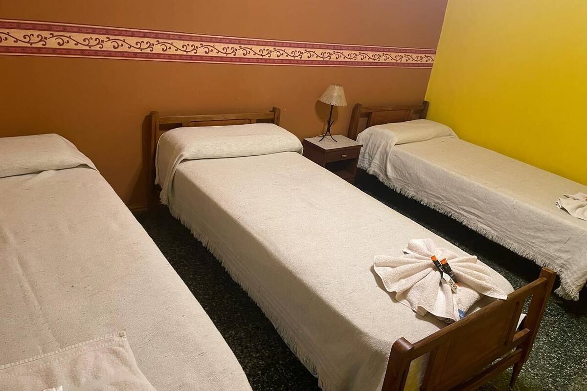 Habitación de hotel con tres camas individuales, una mesita de noche con lámpara y un borde decorativo en la pared.