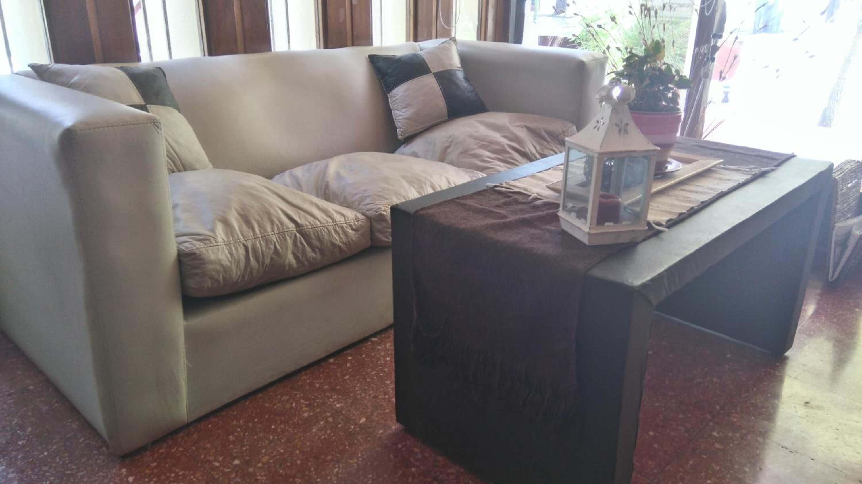 Un sofá color beige con varios cojines, una mesa de centro de madera oscura con un farol de cristal y una planta en maceta al fondo.