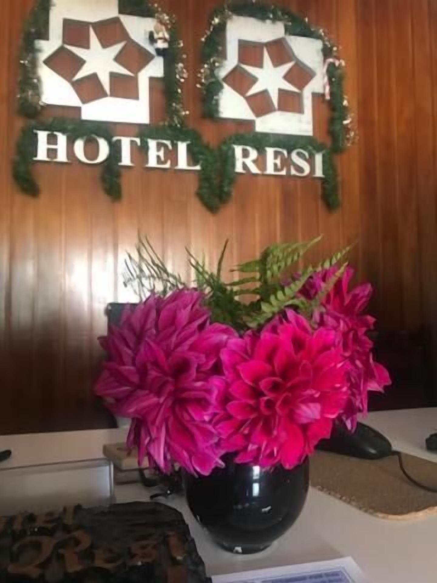 Un comptoir d’accueil d’hôtel avec un vase noir contenant des fleurs roses, des murs recouverts de panneaux en bois et une enseigne indiquant « HÔTEL RESI ».