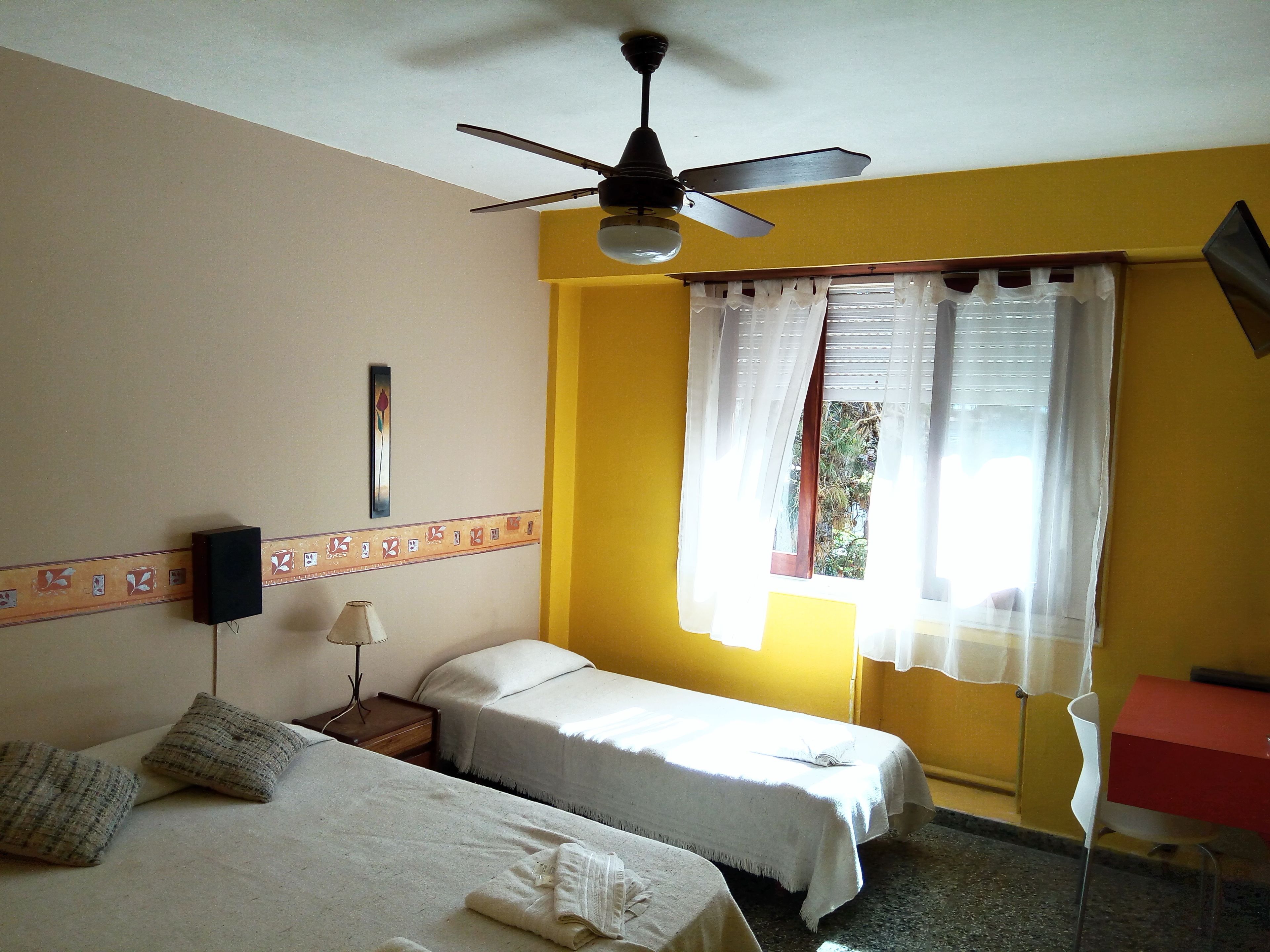 A room with two beds, a ceiling fan, a window with curtains, a wall-mounted TV, and a small table with a lamp.