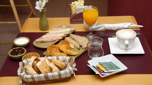 Desayuno con cruasanes, jamón, queso y pan sobre una mesa, acompañado de un vaso de zumo de naranja, una taza de café y un florero con flores.