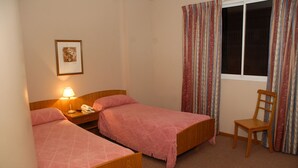 Habitación de hotel con dos camas individuales, una mesita de noche con lámpara, una silla y una ventana con cortinas.