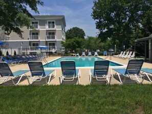 Un espace piscine avec des chaises longues, un bâtiment doté de balcons et un parking clôturé.
