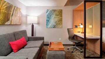 A hotel room with a grey sofa, a red pillow, a wooden desk, a chair, and a large mirror.