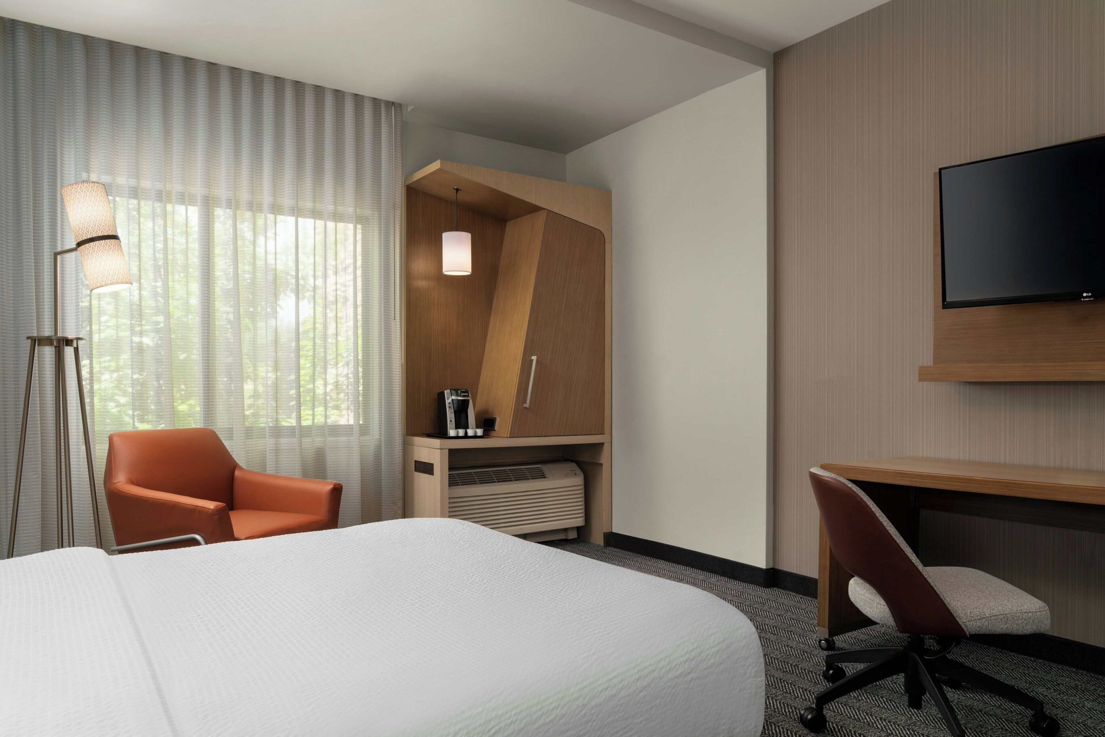 Ein Hotelzimmer mit einem großen Bett, einem Schreibtisch, einem orangefarbenen Sessel, einem an der Wand montierten Fernseher und einem Fenster mit durchsichtigen Vorhängen.