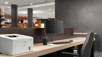 A modern hotel lobby with a reception desk, a large printer, a flat-screen monitor, and a comfortable seating area.
