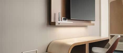 A hotel room with a flat-screen TV mounted on a wooden panel, a desk with a chair, and a built-in heater.