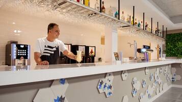 Una zona de bar con un cantinero, una barra con panel de control digital y estanterías con diferentes botellas y vasos.