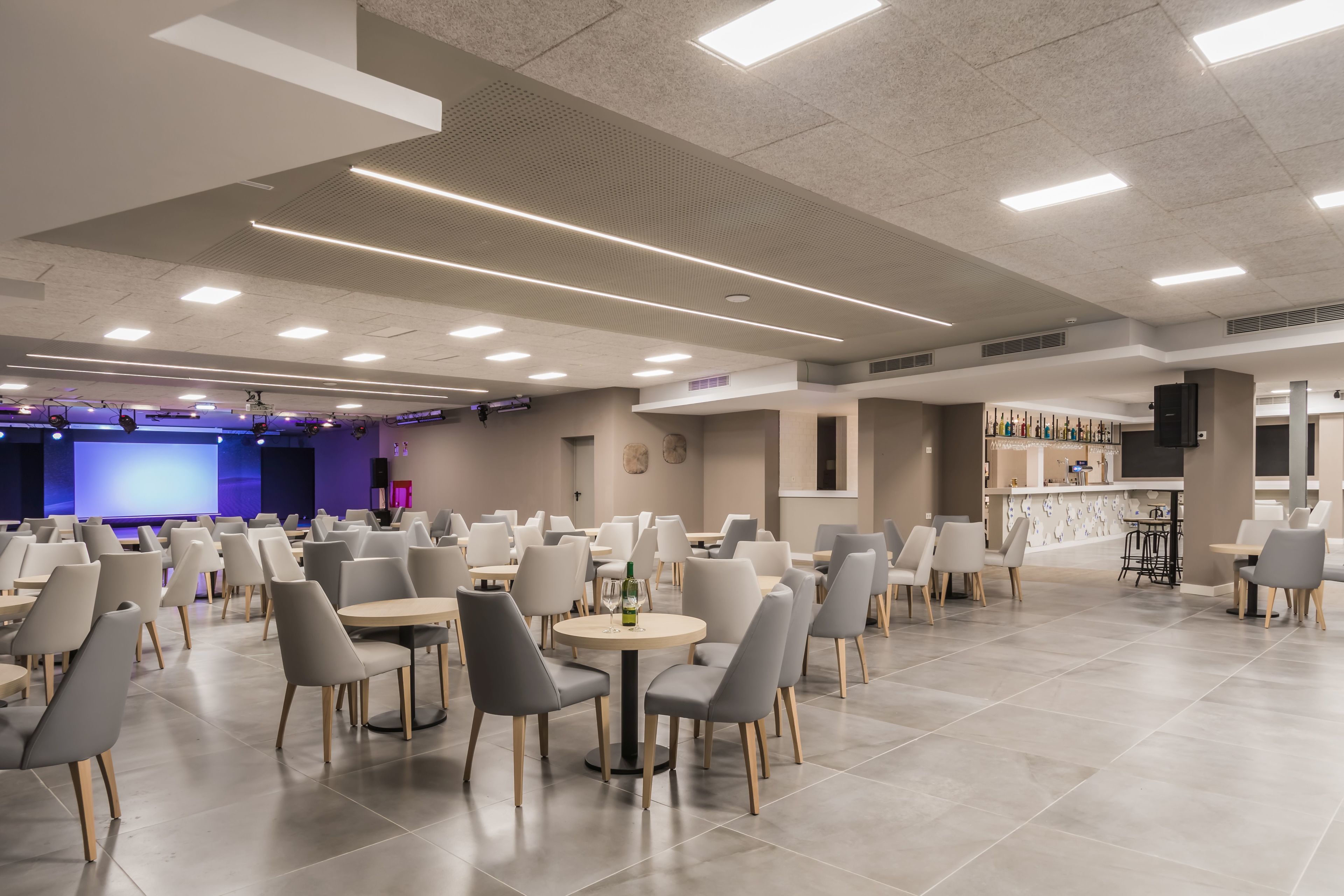 Un comedor moderno con mesas redondas y sillas grises, un escenario con iluminación azul y una zona de bar con botellas.