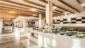 Une cuisine spacieuse avec des poutres en bois, un îlot central sur lequel sont disposés des plats remplis de nourriture, et une personne qui prépare les repas.