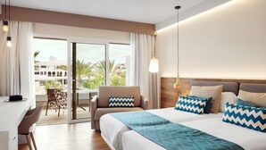 Une chambre d’hôtel avec un grand lit, un bureau et une chaise. Elle dispose d’un balcon avec vue sur les palmiers et les immeubles.