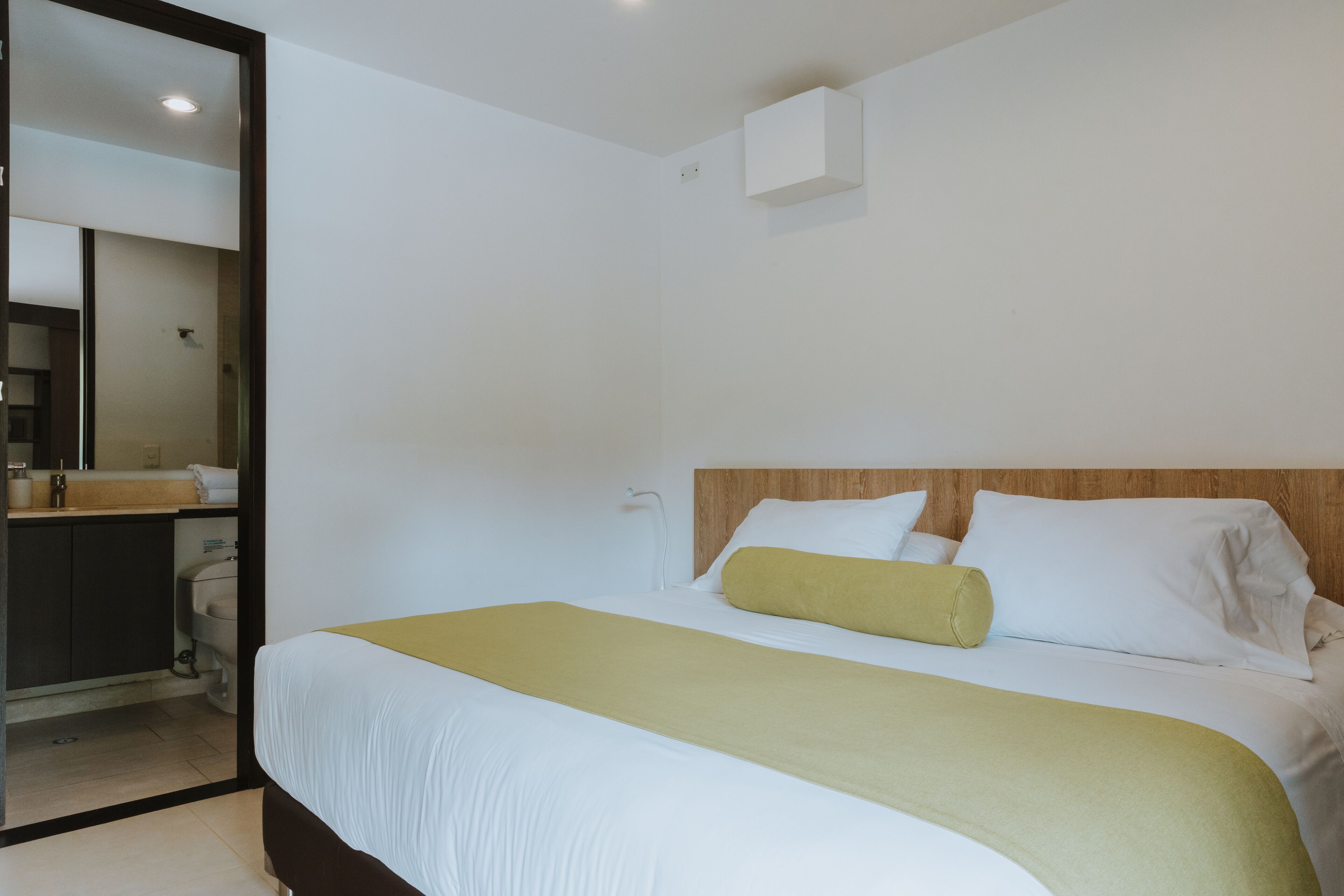 Une chambre d’hôtel avec un lit, une tête de lit en bois, un mur blanc et une petite armoire blanche.