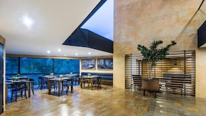 A spacious dining area with multiple tables and chairs, a large window offering a view of the outdoors, and a potted plant in the center. - Travelers Rio Verde Living Suites (Rionegro)