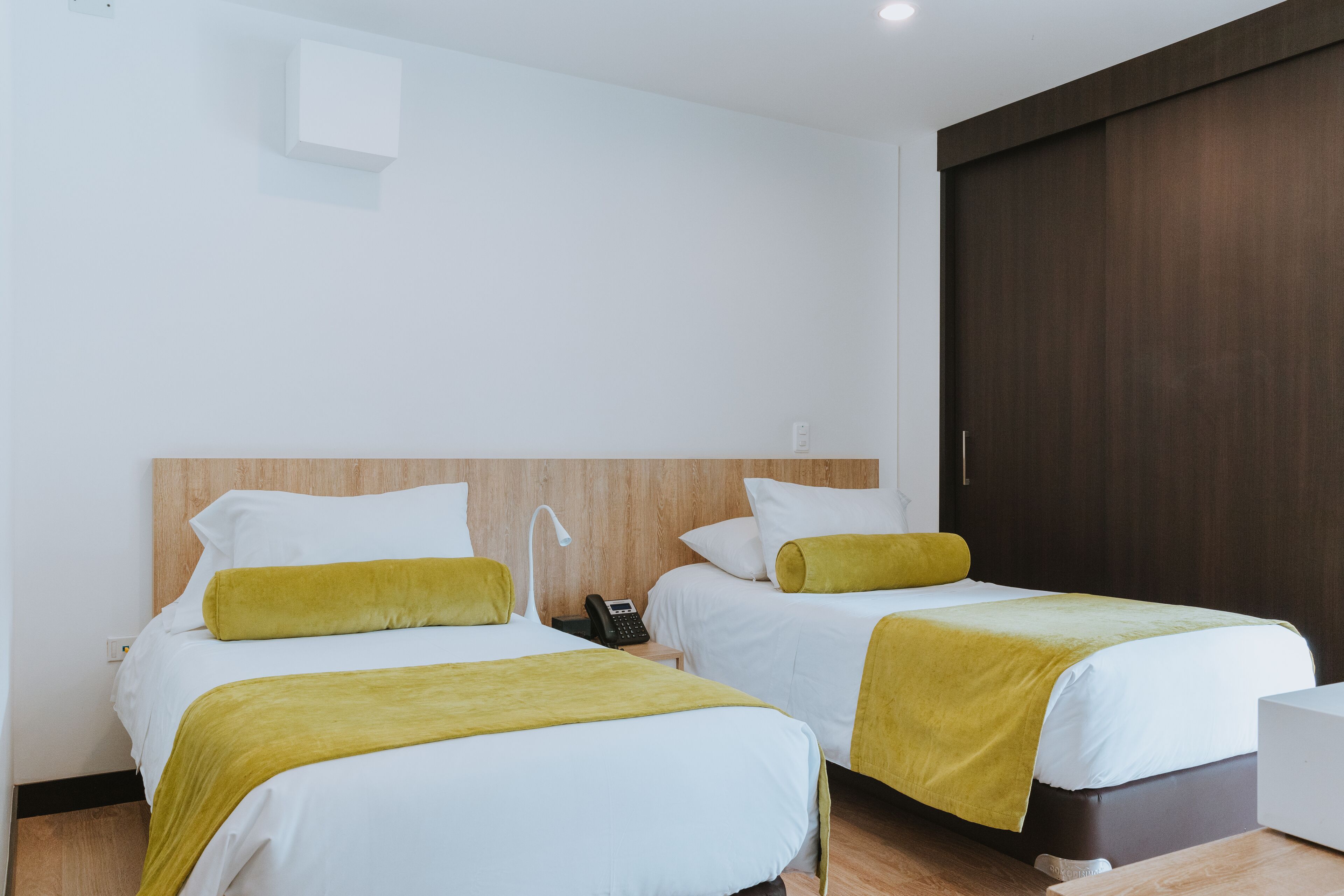 Camera d'albergo con due letti, ciascuno con biancheria bianca e una coperta gialla. È presente una testiera in legno, un comodino con una lampada e un telefono.