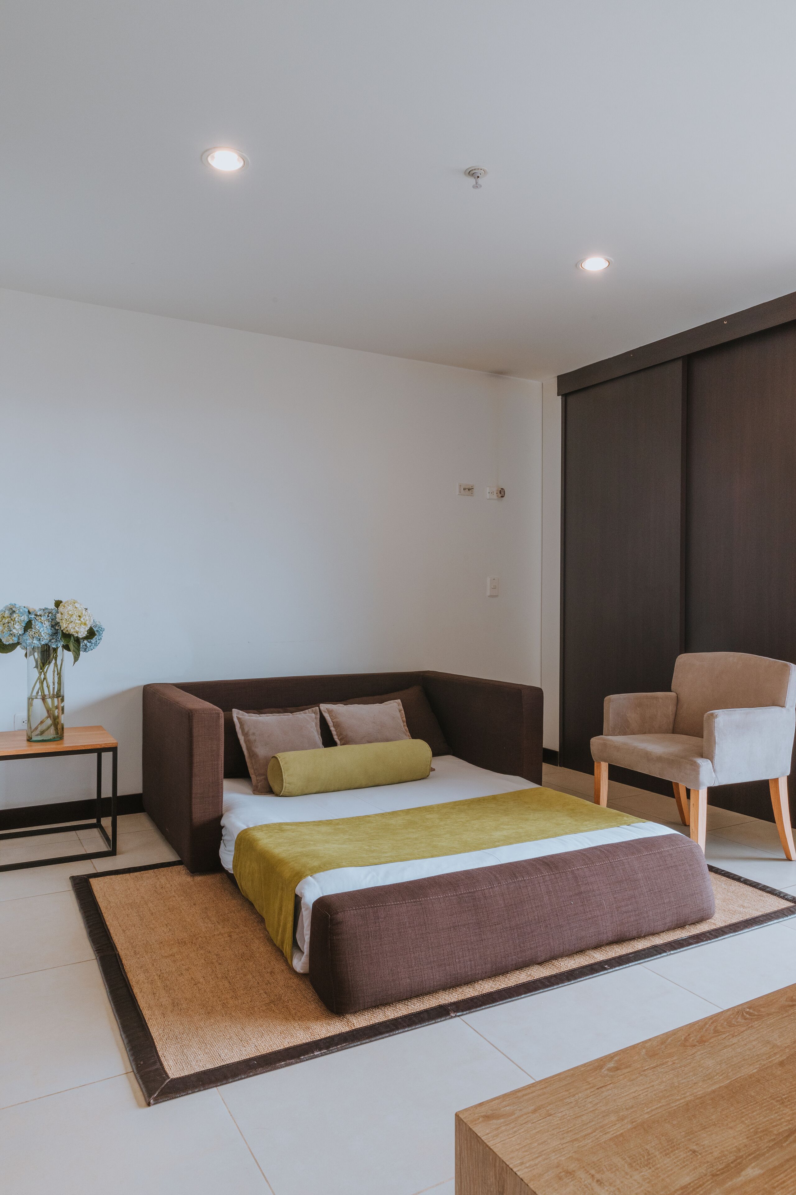 Un dormitorio moderno con una cama, una silla, una mesita y un jarrón de flores.
