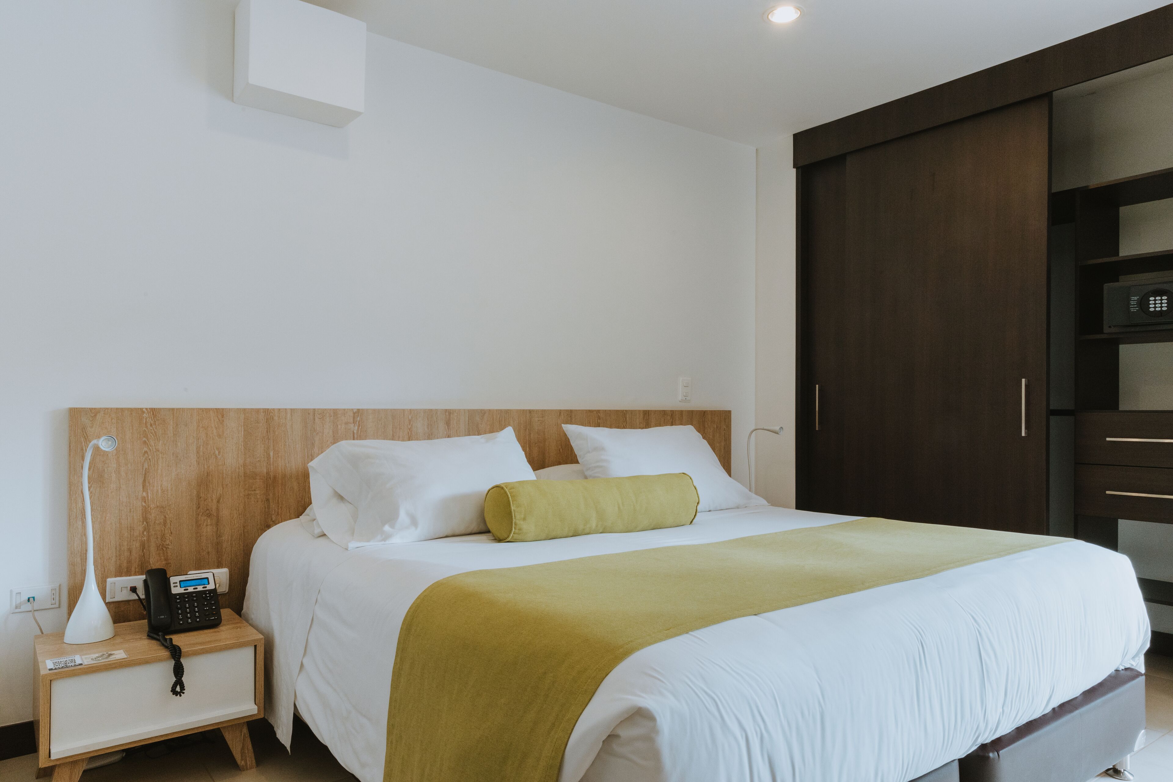 Une chambre d’hôtel avec une tête de lit en bois, un lit avec une literie blanche et un plaid vert, une table de chevet avec une lampe et un téléphone, et une armoire à la finition sombre.