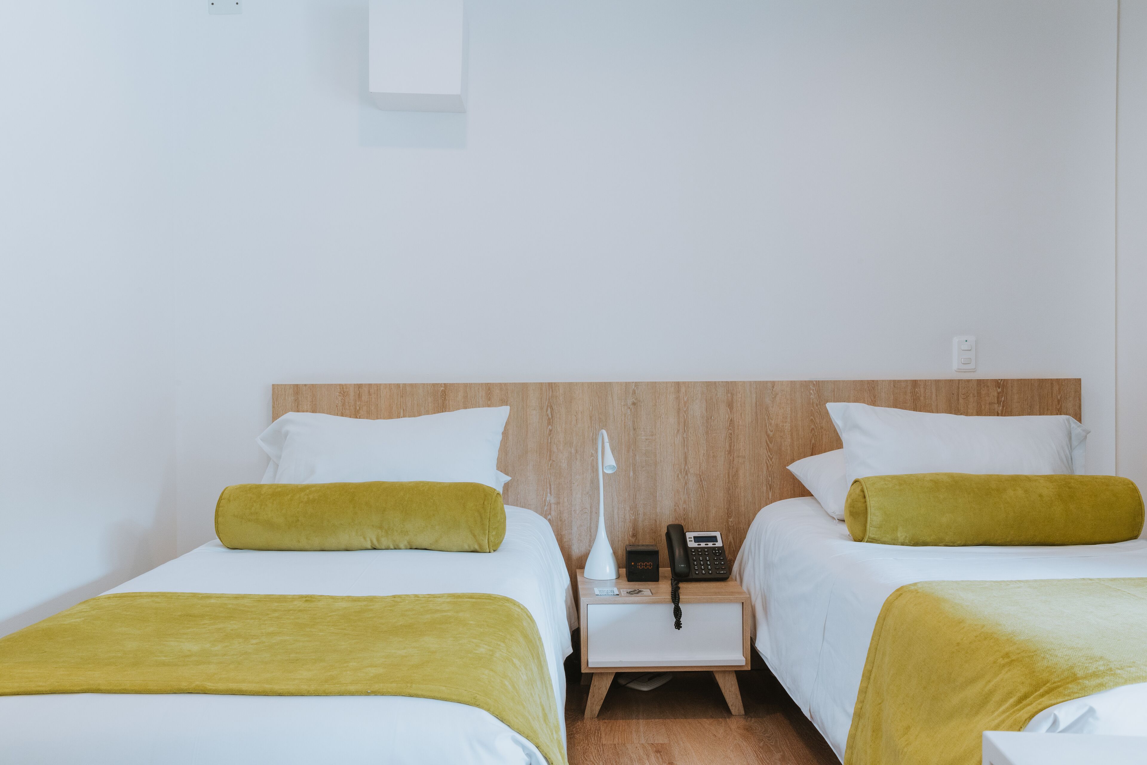 A hotel room with two beds, each with white bedding and a yellow throw blanket. There is a bedside table with a lamp, a phone, and a small clock. The room has a wooden headboard and white walls.
