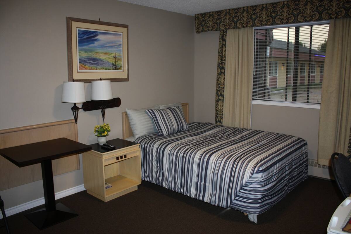A hotel room with a bed, a desk, a chair, a lamp, a painting on the wall, and a window with curtains.