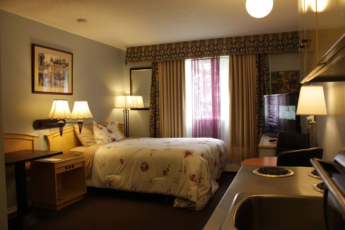 A hotel room with a bed, two bedside lamps, a television, a desk with a computer, and a window with curtains.