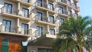 Un hotel de varios pisos con balcones, un coche estacionado adelante y un área ajardinada con plantas.