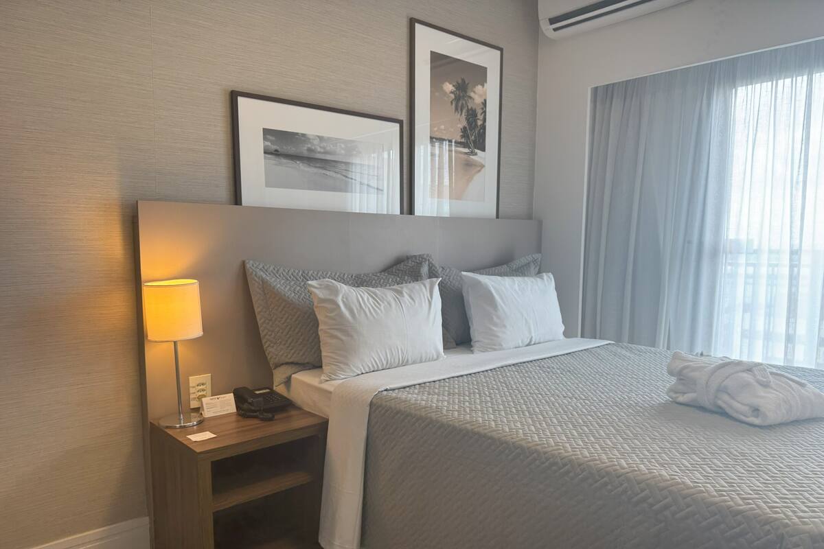 A neatly made bed with a grey quilt, a bedside lamp, a phone, and a small wooden nightstand.