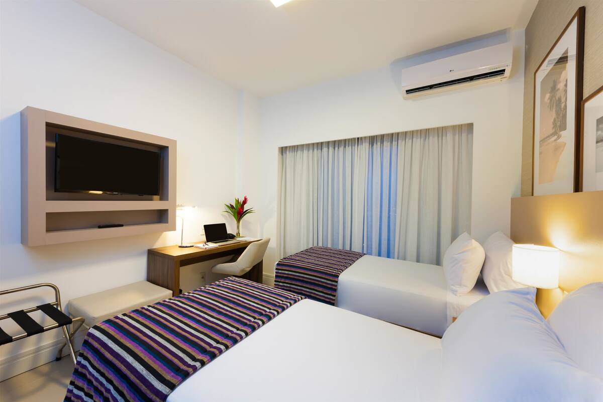 A hotel room with two beds, a desk, a chair, a TV, and an air conditioning unit.