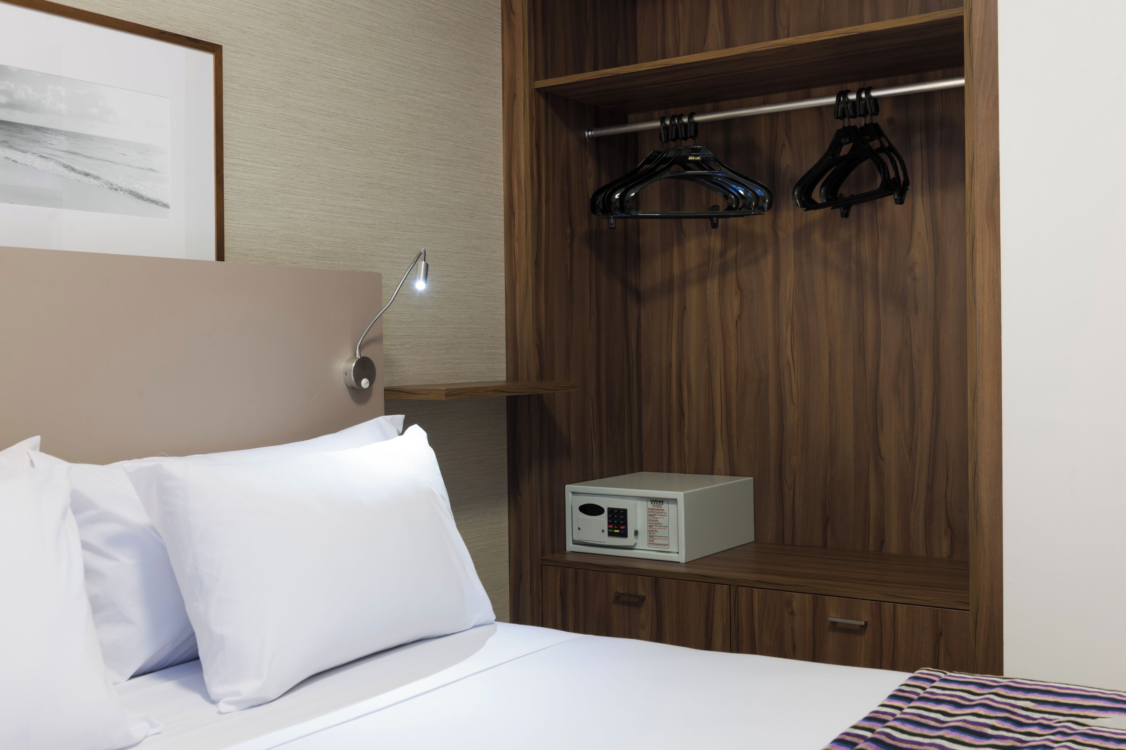 Camera d'albergo con un letto, un comodino con una lampada, un armadio con grucce e un piccolo elettrodomestico sullo scaffale.