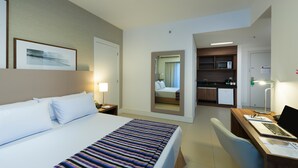 Una habitación de hotel con una cama, un escritorio con una computadora portátil, una silla, un espejo, una puerta que da a un baño y una zona de kitchenette.
