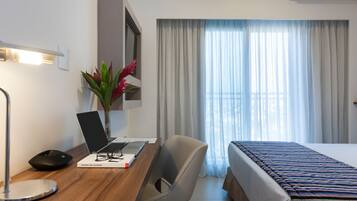A hotel room with a wooden desk, a laptop, a chair, a bedside table, a bed, a window with curtains, and a potted plant.