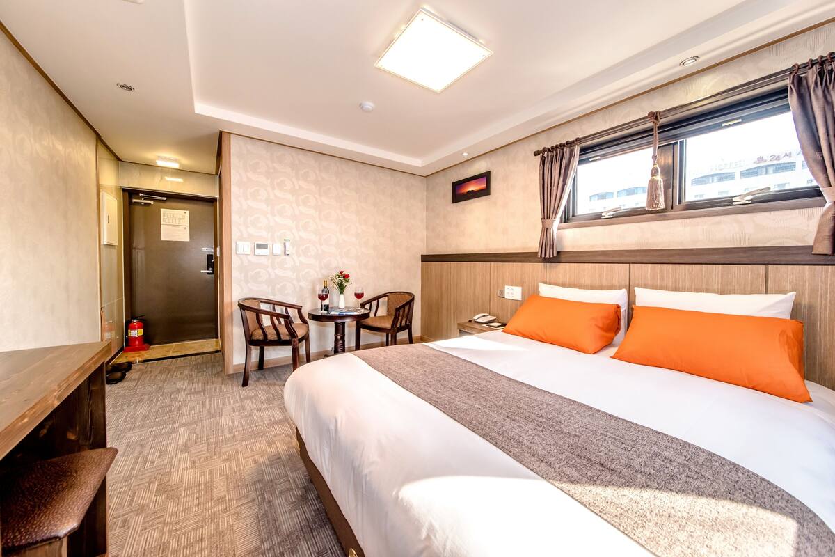 Une chambre d’hôtel avec un grand lit, un bureau, une chaise, une petite table avec un vase et des verres à vin, et une fenêtre avec des rideaux.
