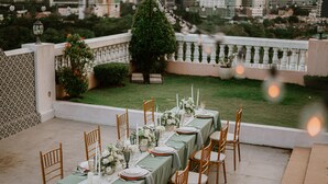 A long dining table set outdoors with chairs, surrounded by greenery and a cityscape in the background.