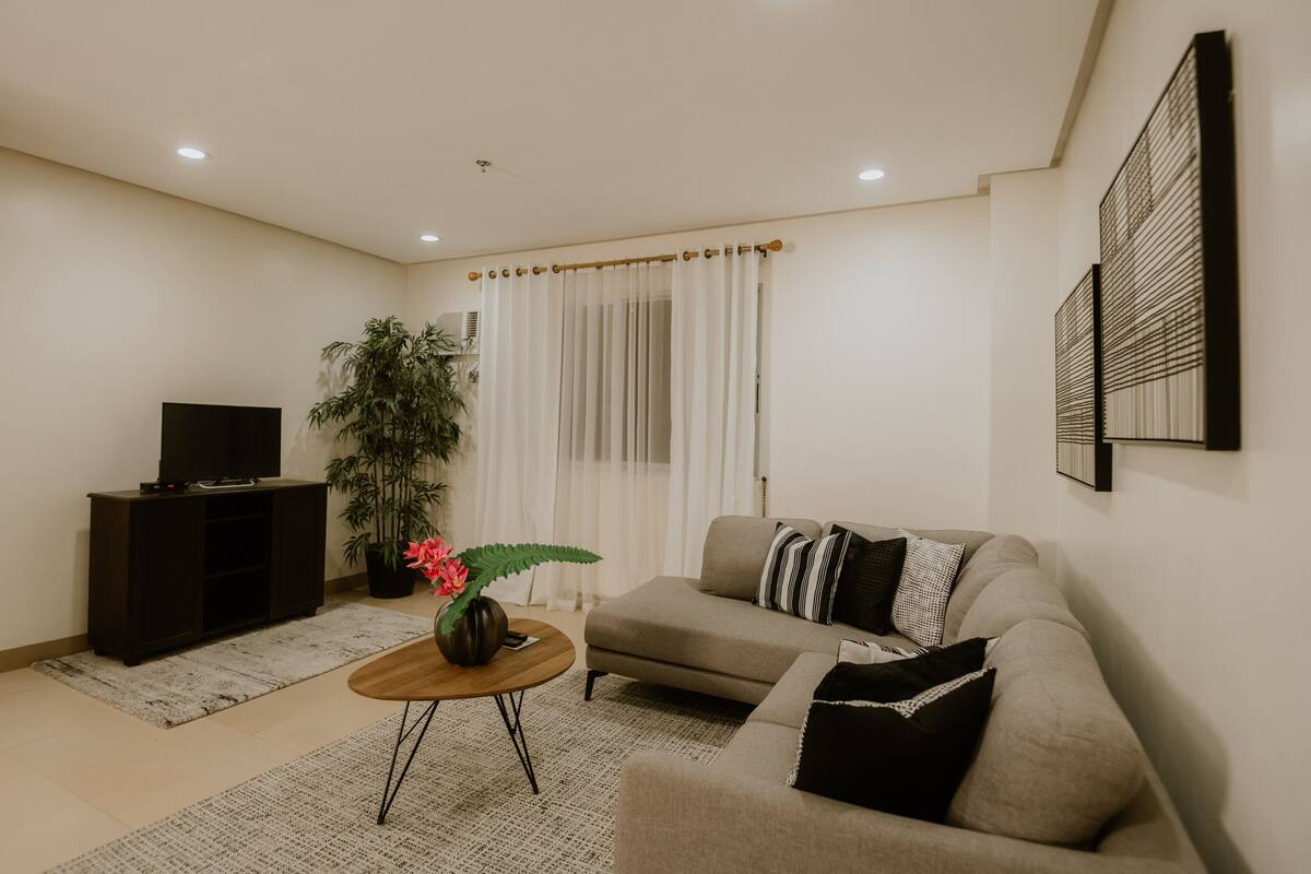 Sala de estar con sofá, una mesa de centro, televisor sobre un mueble, planta en maceta y una cortina.