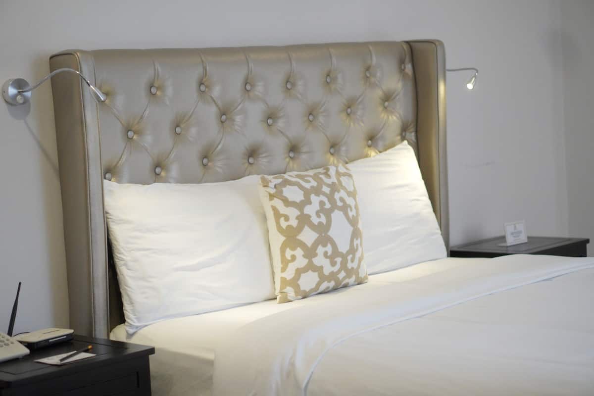 Una cama bien hecha con cabecera capitonada, una mesita de noche con lámpara y un pequeño letrero en la mesita.