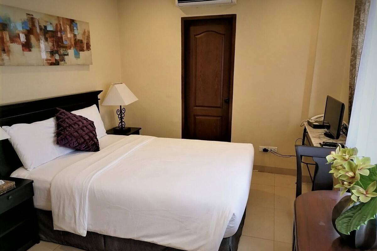Una cama bien hecha con sábanas blancas, un cabecero oscuro y una almohada morada. Hay una mesita de noche pequeña con una lámpara y un aire acondicionado instalado en la pared encima de la cama. Se ve una puerta de madera al fondo, y un escritorio con una computadora y una planta a la derecha.