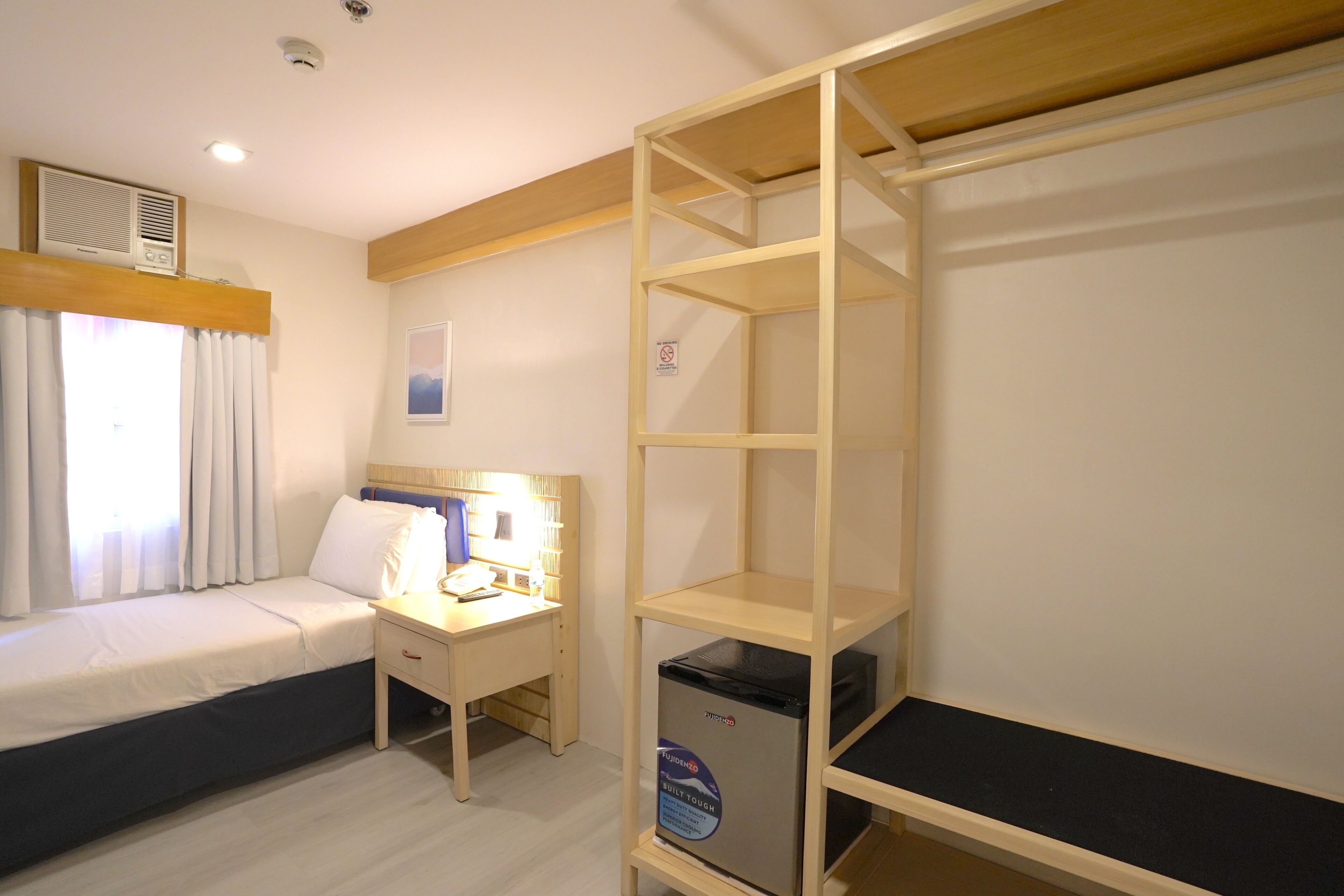 A hotel room with a bed, a desk with a computer, a small refrigerator, and a bookshelf.