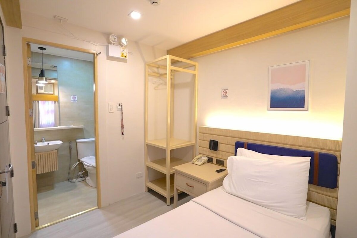 A hotel room with a bed, bedside table, a phone, a shelf, a window, and a view of a bathroom.