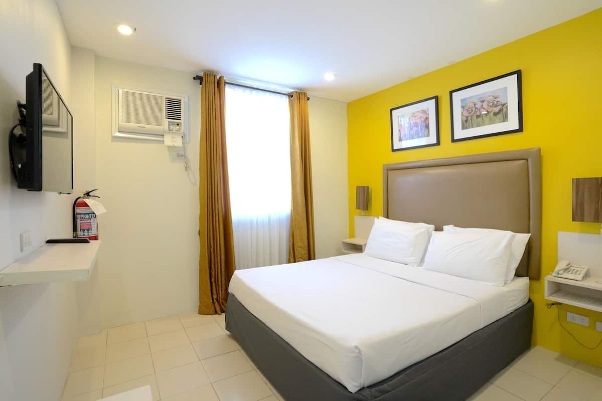 Una habitación de hotel con cama, televisión, extintor, ventana con cortinas y una pared con dos cuadros enmarcados.