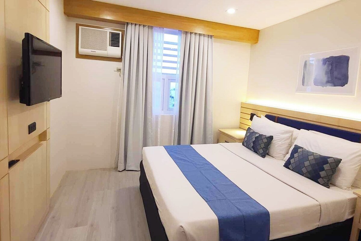 A hotel room with a bed, a TV, an air conditioning unit, a window with curtains, and a wall-mounted artwork.