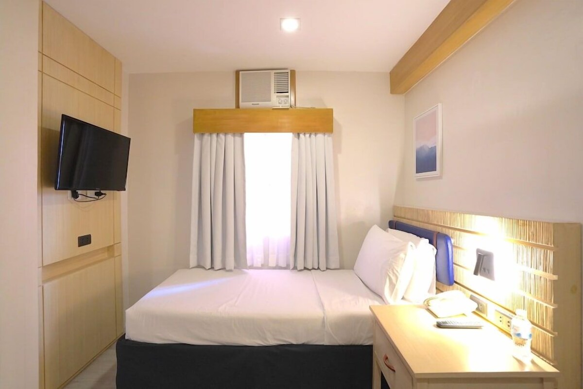 A hotel room with a bed, a TV mounted on the wall, a bedside table with a lamp, and an air conditioning unit above the window.