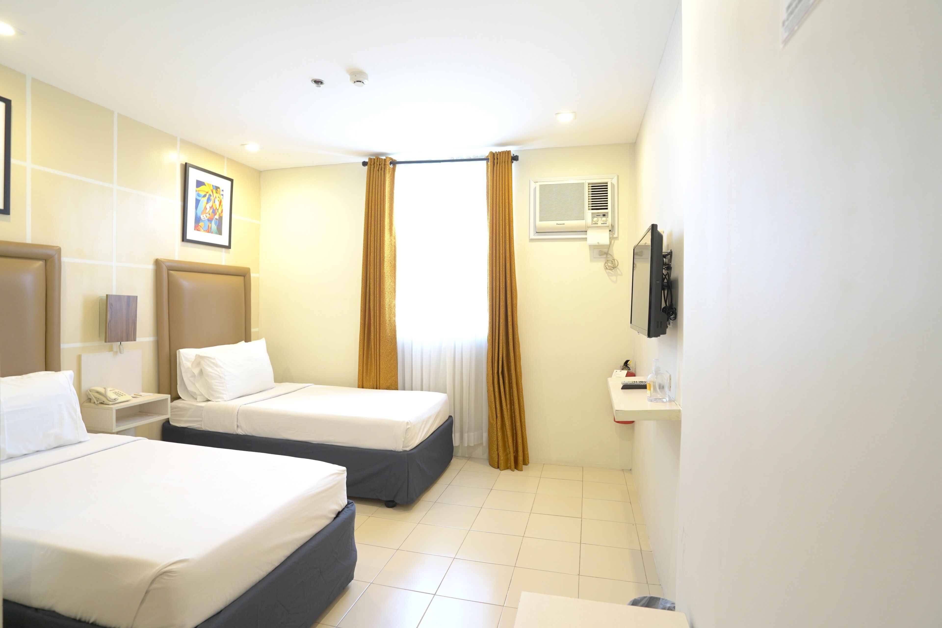 A hotel room with two beds, a bedside table with a phone, a wall-mounted TV, and a window with curtains.