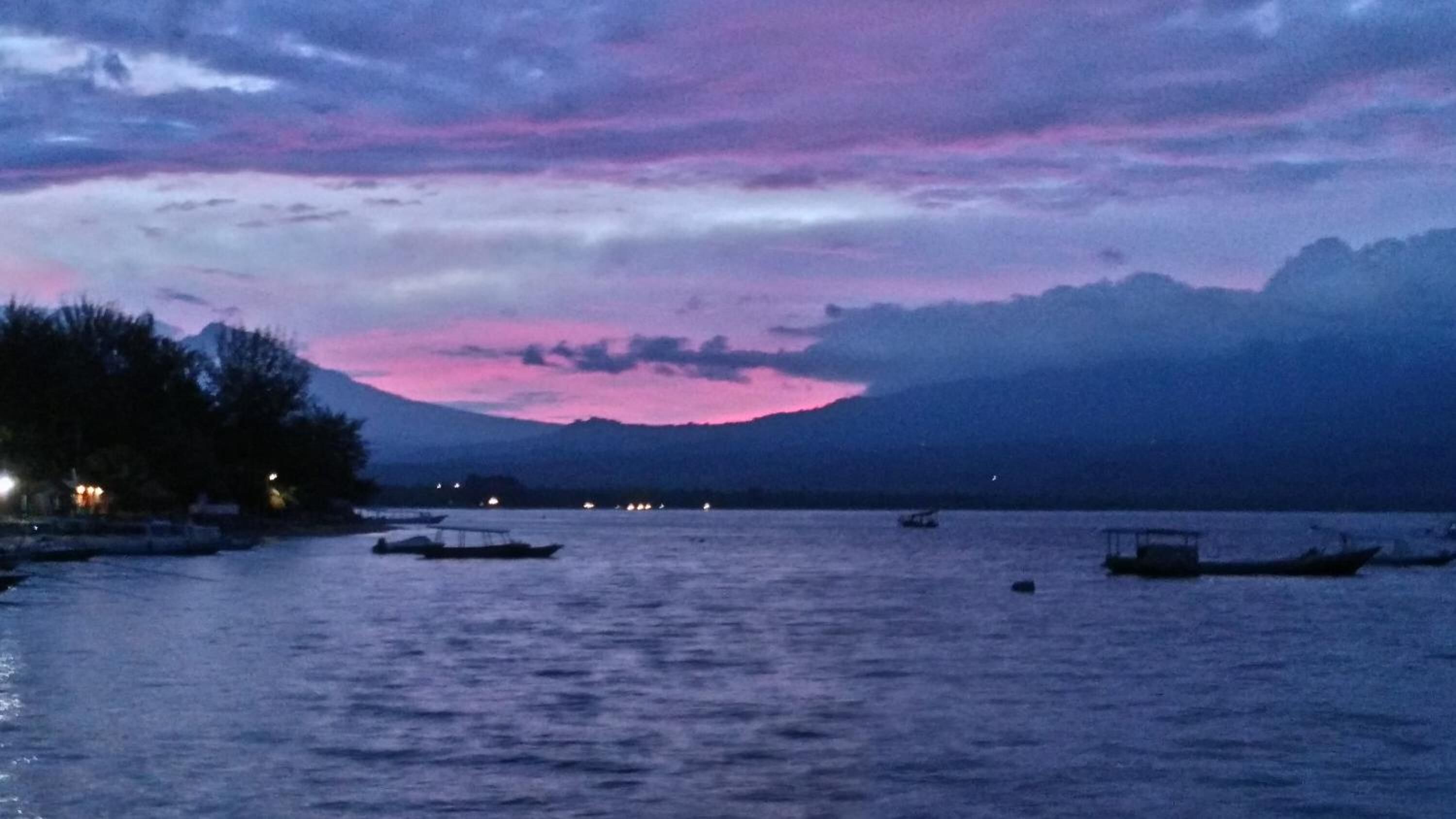 A calm lake at dusk with boats and a mountain in the background.