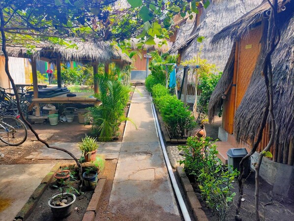 A narrow pathway flanked by thatched-roof huts, potted plants, and a bicycle.