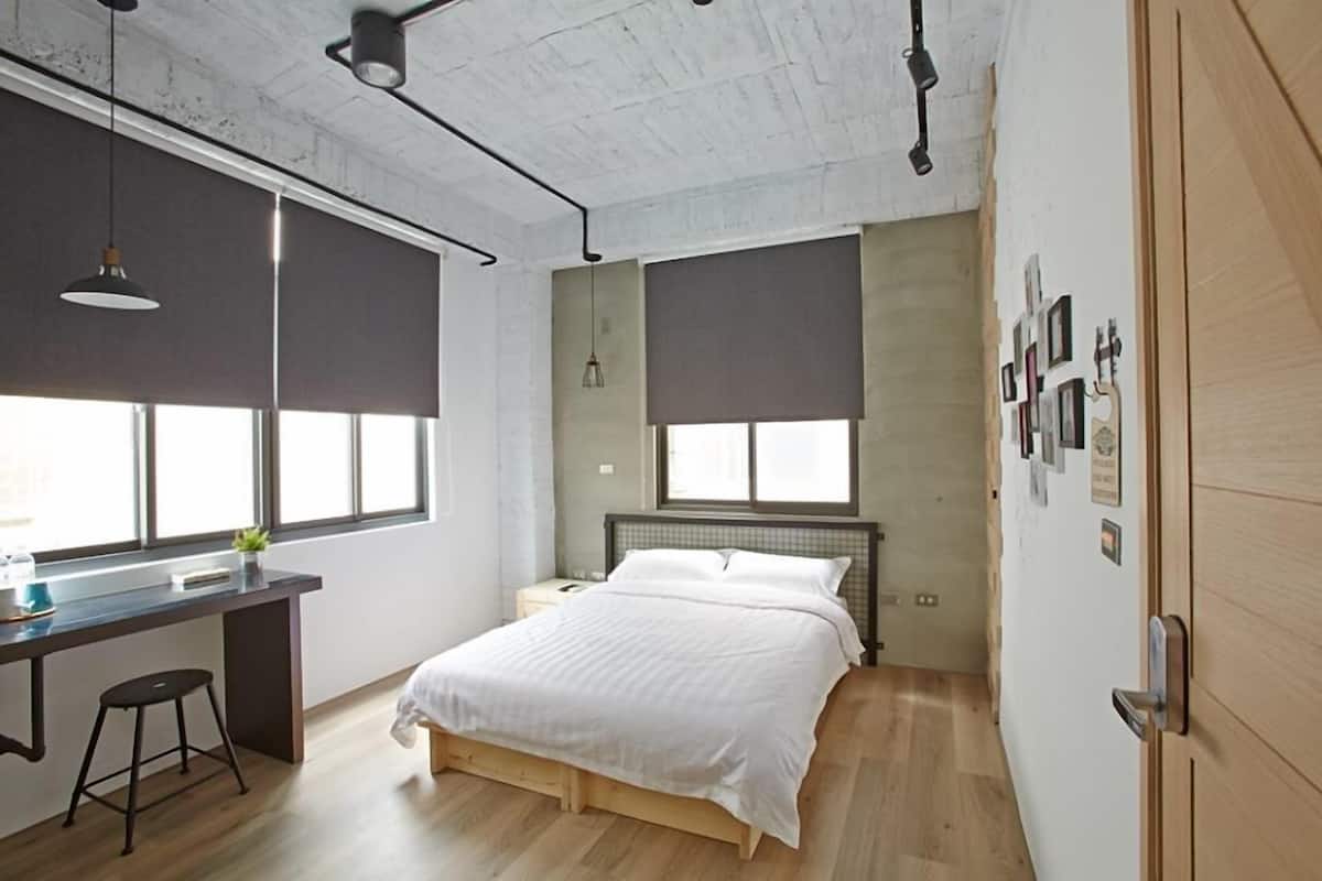 Un dormitorio moderno con una cama, un escritorio y una silla. Hay dos ventanas con persianas, una lámpara en el techo y una puerta con pomo.