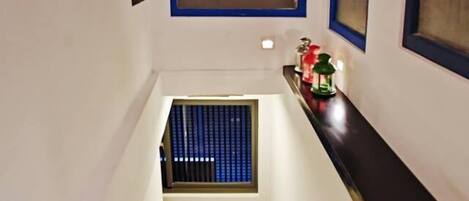 Una escalera con escalones de madera y barandilla oscura, que conduce a una plataforma con un ventanal pequeño y un estante decorativo.