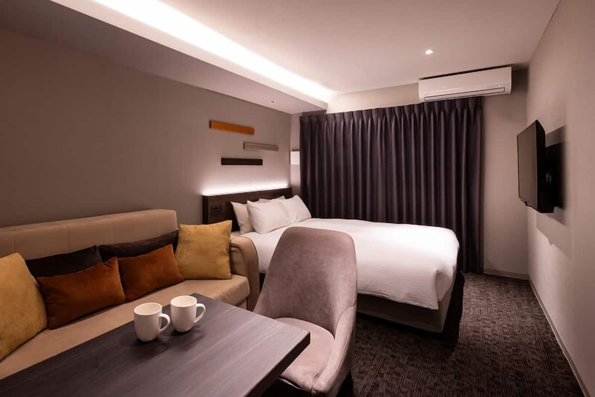 A hotel room with a bed, a sofa, a table with a coffee cup, and an air conditioning unit.