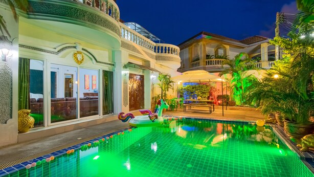 A swimming pool with a green-lit edge, surrounded by a well-maintained garden and a large, multi-story building with balconies. - Ratana Villa - Pattaya Holiday House Walking Street (Pattaya)