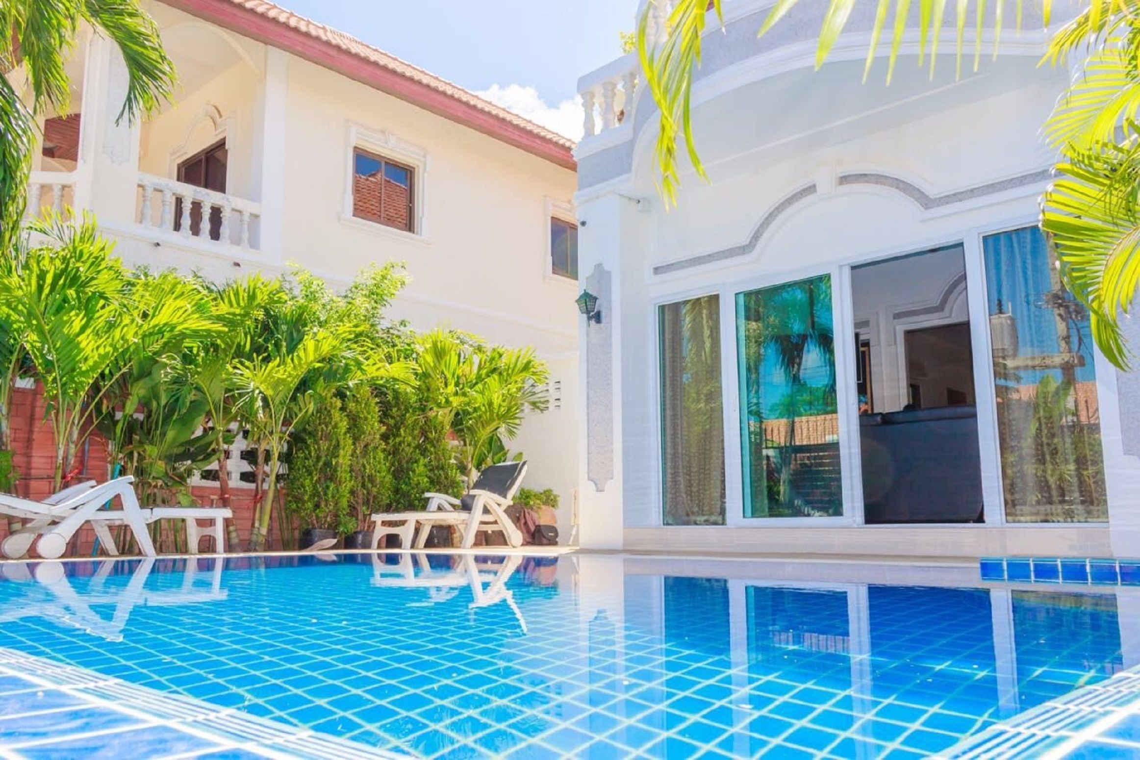 A swimming pool with tiled flooring, surrounded by lush greenery and white-framed windows.
